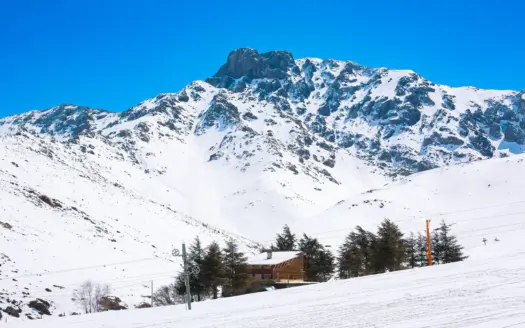 Laderas nevadas, un pilón de telesilla y chalés bajo un pico del Alto Atlas